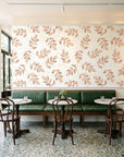 Verdant Scatter Terracotta tossed leaf wallpaper above a green leather banquette in a sunlit neighborhood cafe