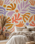 Wallpaper featuring cheerful, abstract botanical elements in soft coral, ochre, lavender, and peach hues. The hand-drawn design rests upon a warm cream base.