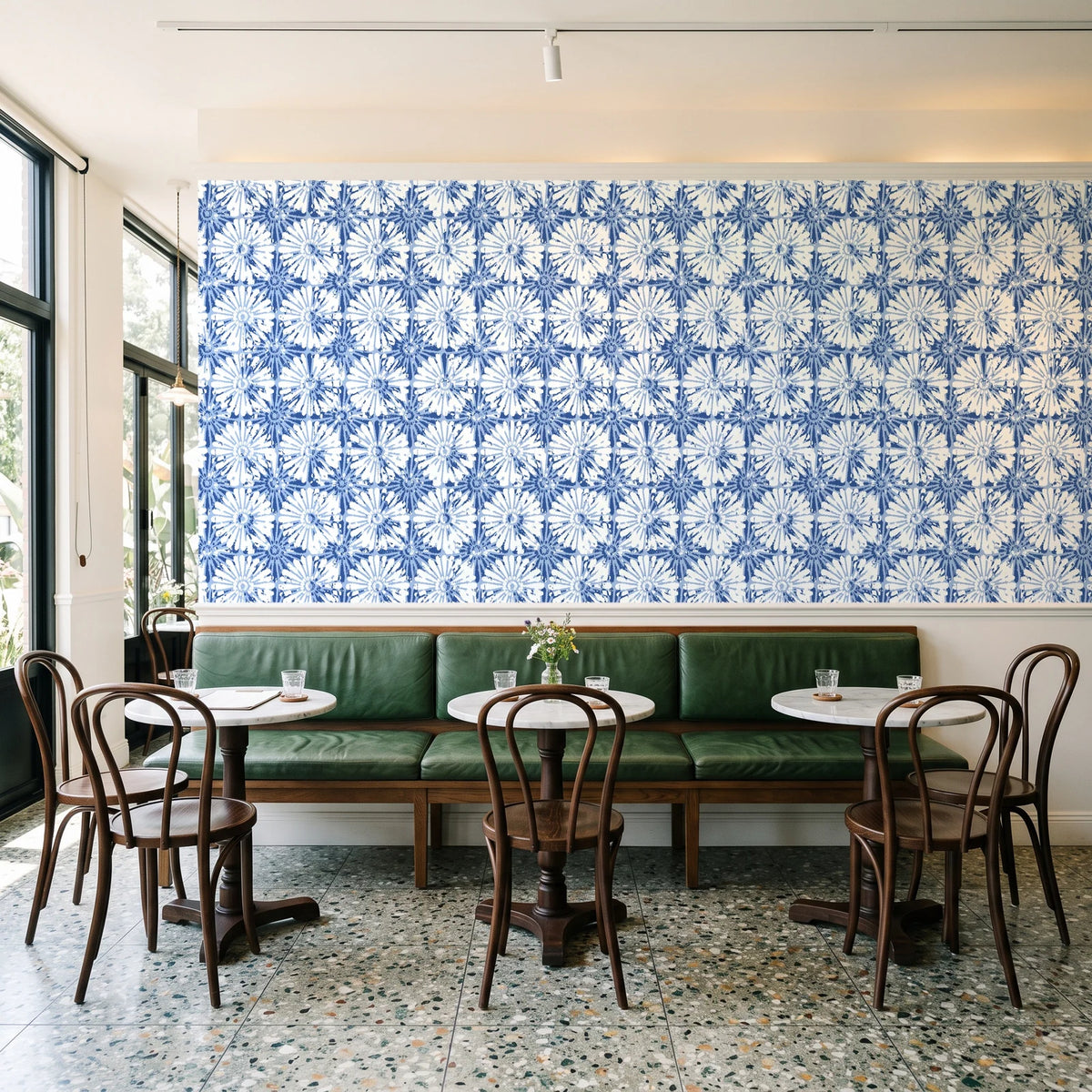 Solar Ritual Indigo floral wallpaper above a green leather banquette in a sunlit neighborhood cafe