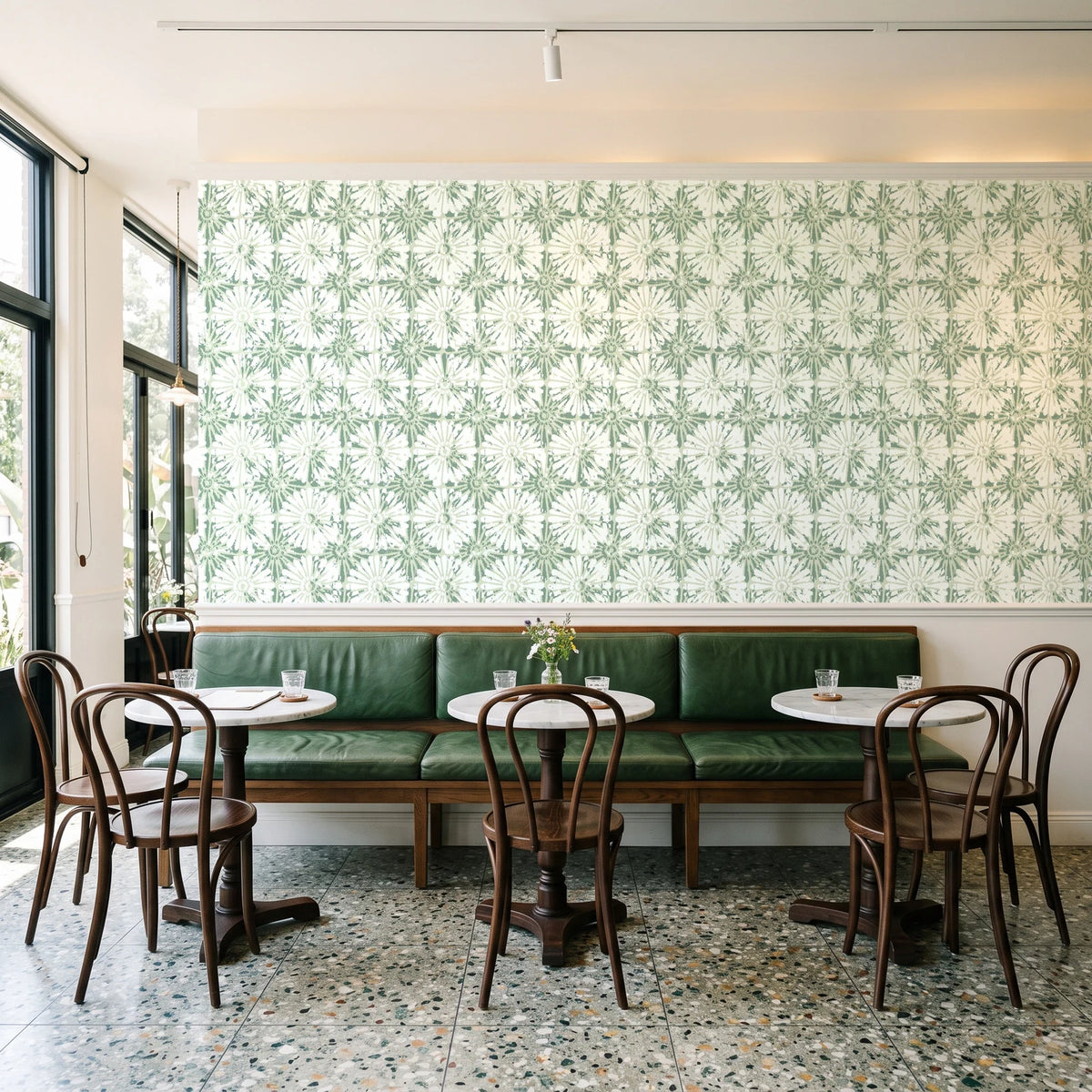 Solar Ritual Sage floral wallpaper above a green leather banquette in a sunlit neighborhood cafe