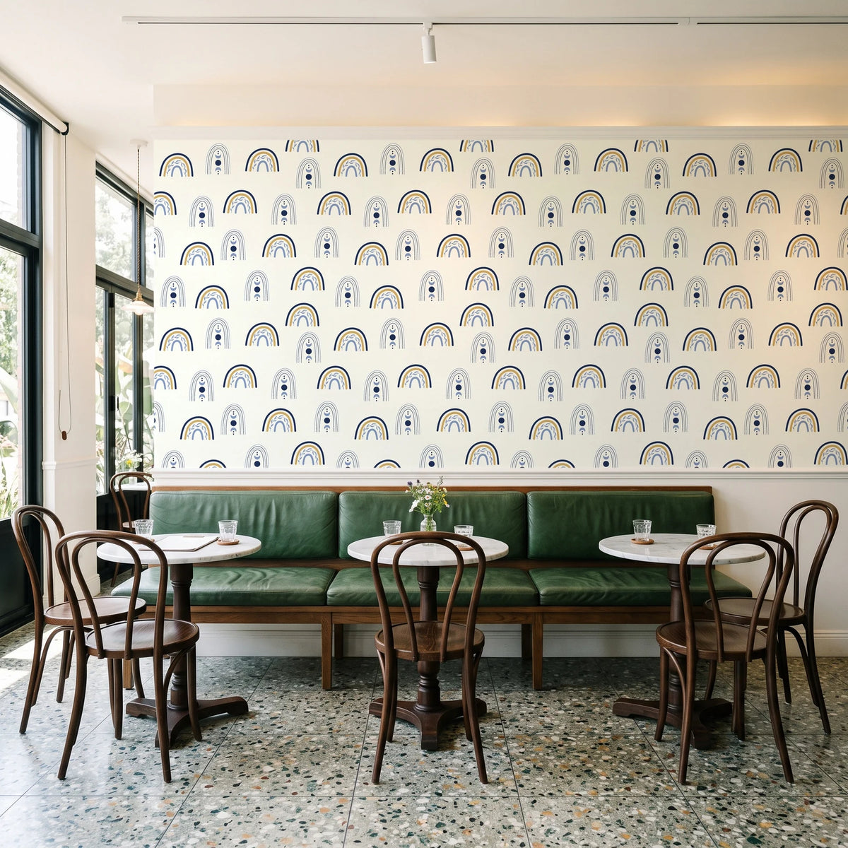 Solstice Rite Midnight boho rainbow wallpaper above a green leather banquette in a sunlit neighborhood cafe