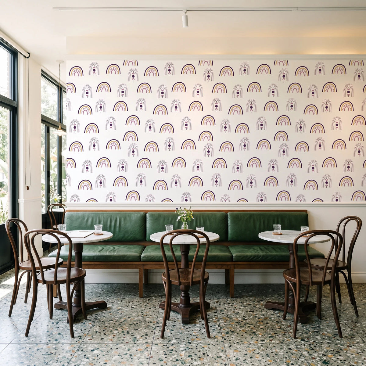 Solstice Rite Plum boho rainbow wallpaper above a green leather banquette in a sunlit neighborhood cafe